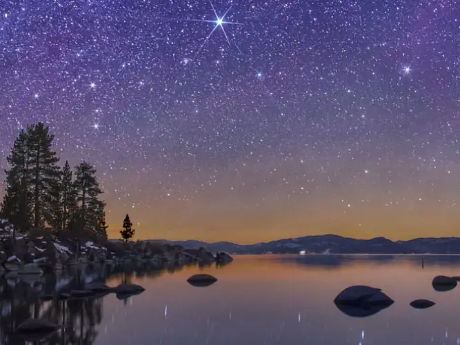 a starry night sky over a lake with trees and rocks on the shore